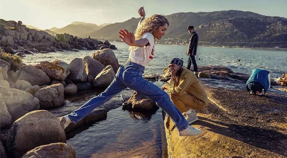 Woman jumps a stream on a rocky coast near the ocean with mountains in background. People are relaxing nearby in the sunshine.