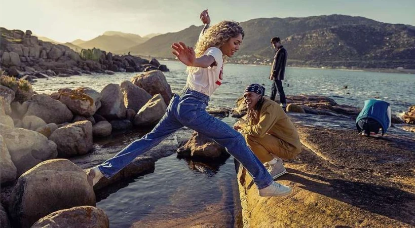 Woman jumps a stream on a rocky coast near the ocean with mountains in background. People are relaxing nearby in the sunshine.