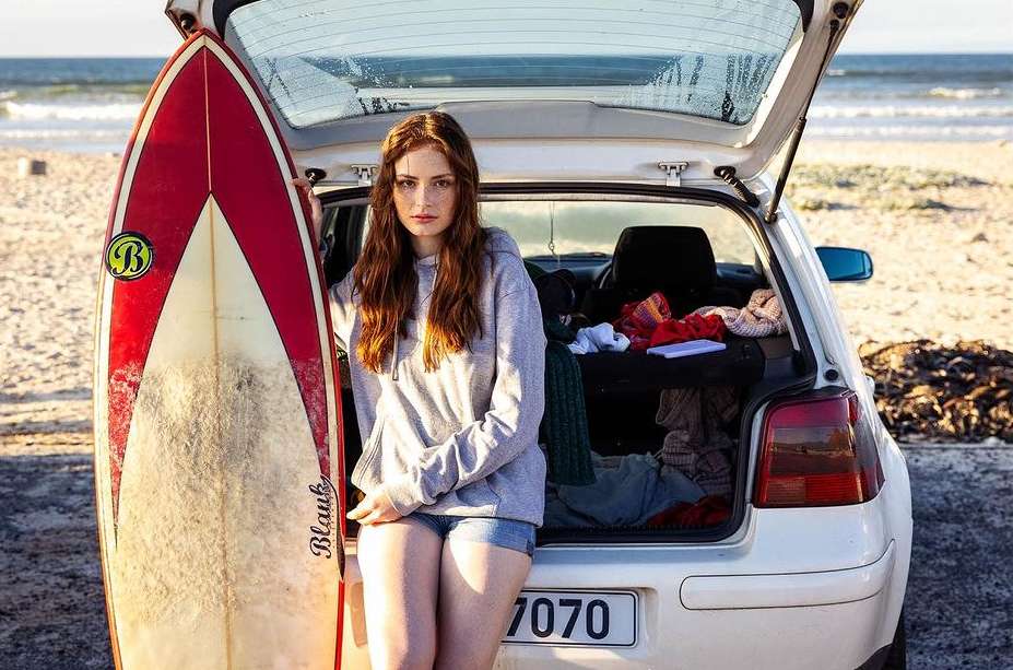 A woman in casual clothes stands next to a surfboard, leaning against a white car with its trunk open on a sandy beach.
