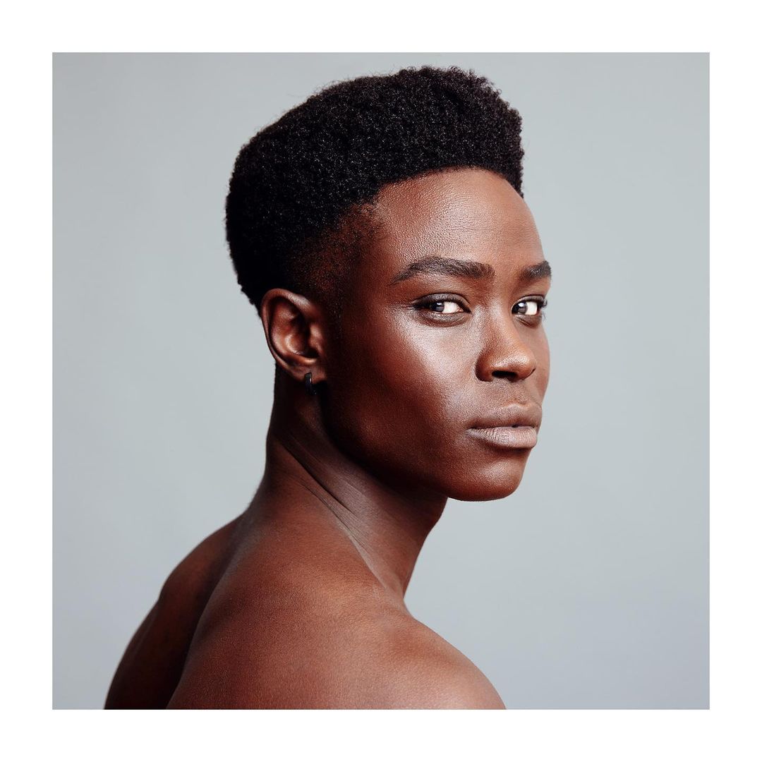 Portrait of a young adult Black man with a dark complexion and short natural hair, wearing subtle makeup, captured in a studio setting.