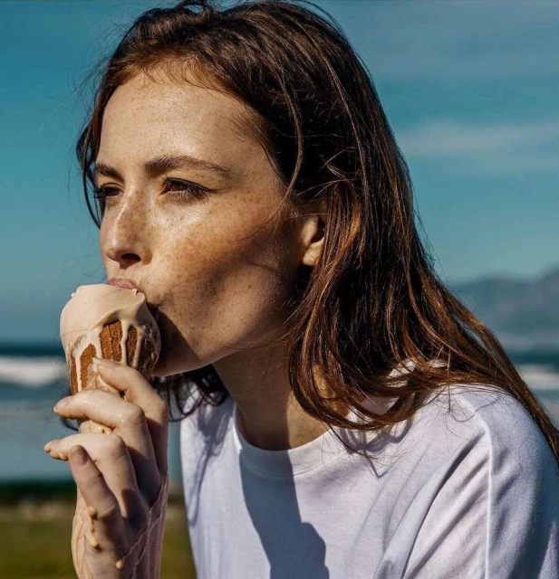 Young woman with freckles enjoying an ice cream cone on a sunny day, ocean and dunes in the background, shallow depth of field.