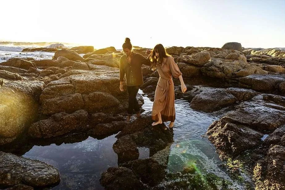A couple walks barefoot on rocky coast at sunset with ocean waves. The woman wears a brown maxi dress, the man a shirt and dark pants.
