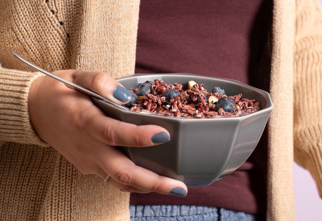 Close-up of a gray bowl filled with cereal, blueberries, and milk, held by a woman in a cream-colored sweater, showcasing a healthy food choice.