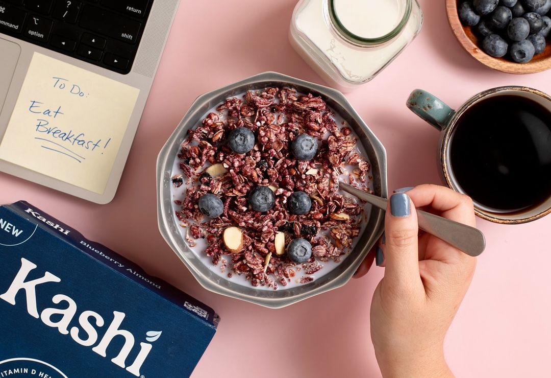 Overhead shot of a breakfast bowl with cereal and blueberries, a laptop, a coffee cup, and a Kashi cereal box on a pink table.