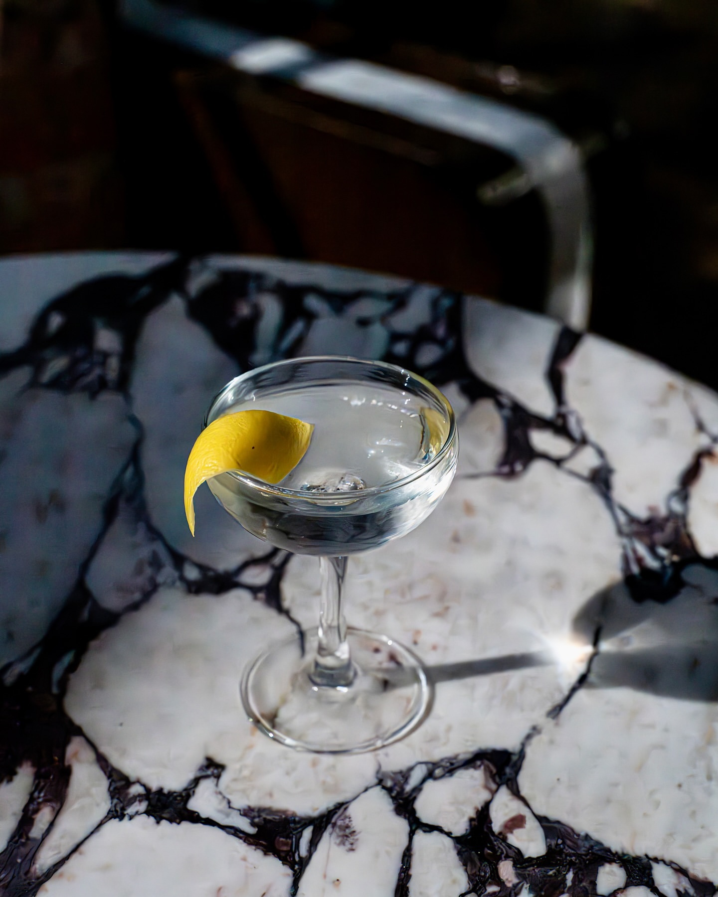 A coupe glass filled with a clear beverage and garnished with a lemon twist sits on a marble table with a bar in the background.