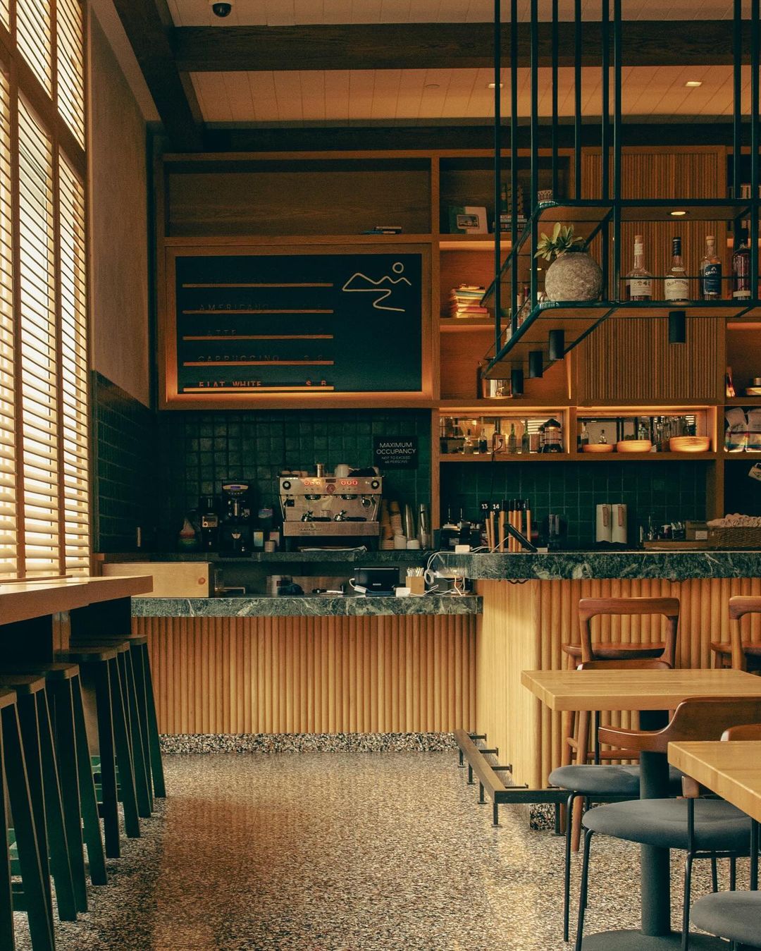 Interior view of a stylish coffee shop featuring a coffee machine, tables, chairs, and a terrazzo floor. Dark green and wood accents.