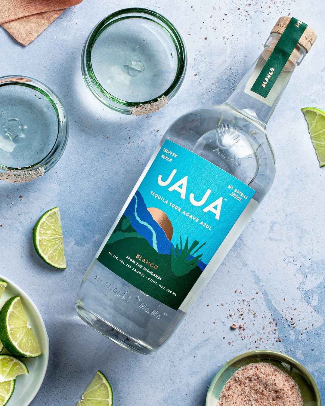 JAJA Tequila bottle with lime wedges and salt-rimmed glasses set on a light blue surface in a visually appealing food and beverage product shot.