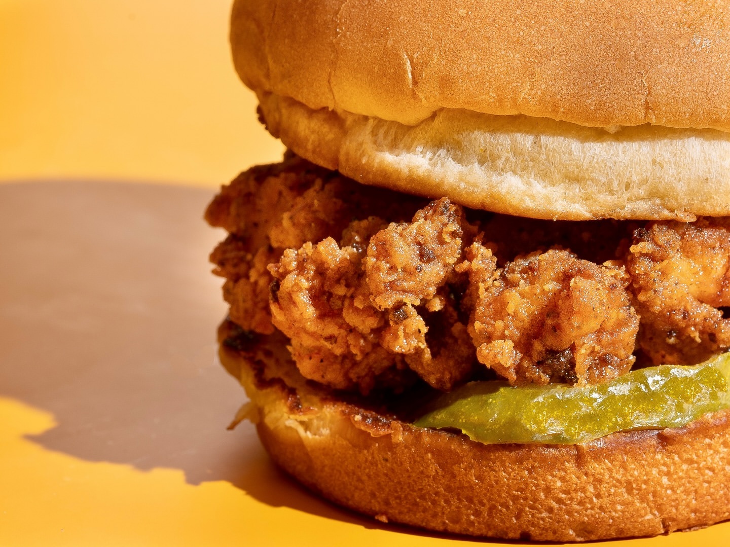 Close-up of a chicken sandwich with a toasted bun, fried chicken, and pickles, set against a sunny yellow background.