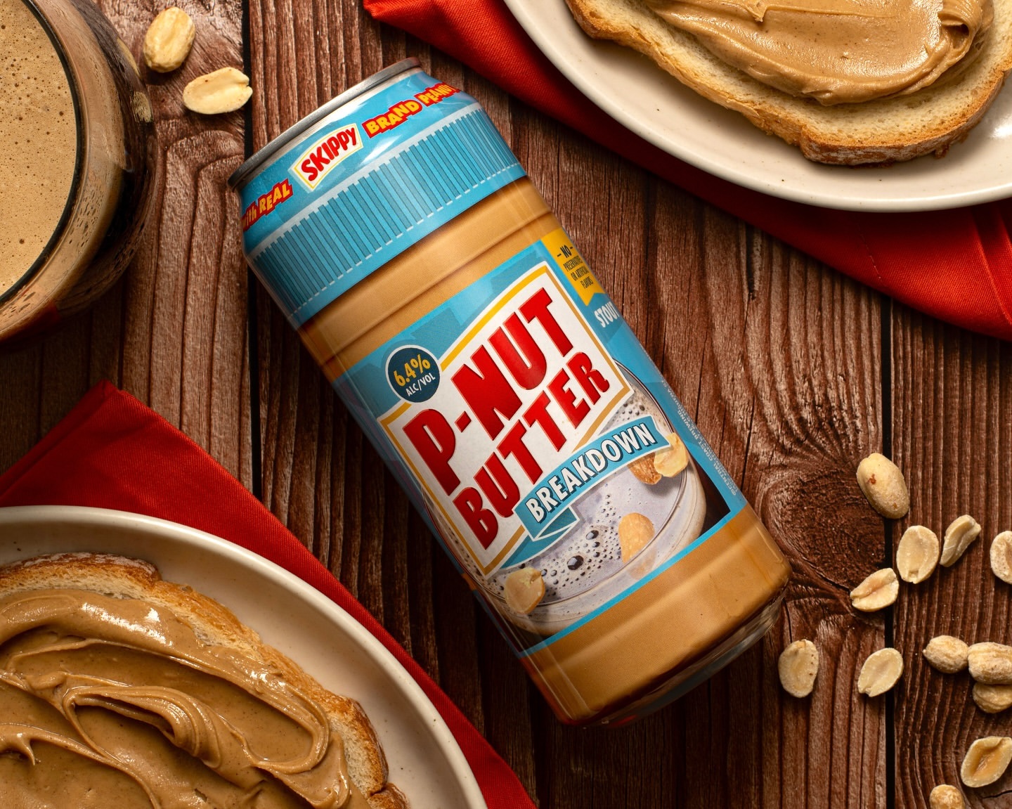 Overhead shot of a can of P-Nut Butter Breakdown Stout beside a red napkin, peanuts, and bread with peanut butter on a wooden table.