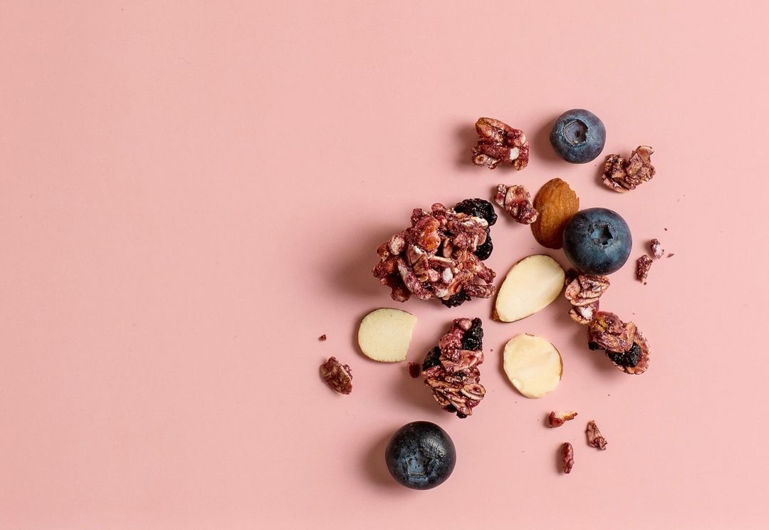 Overhead shot of granola clusters, almonds, and blueberries on a pink background, capturing a healthy food flat lay composition.