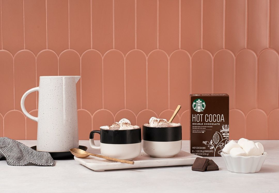 Hot cocoa drinks with marshmallows and chocolate pieces presented with a ceramic pitcher, mugs, dishware, and a box of cocoa mix.