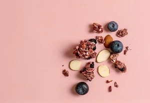 Top-down view of granola clusters, whole blueberries, and sliced almonds scattered on a solid pink background