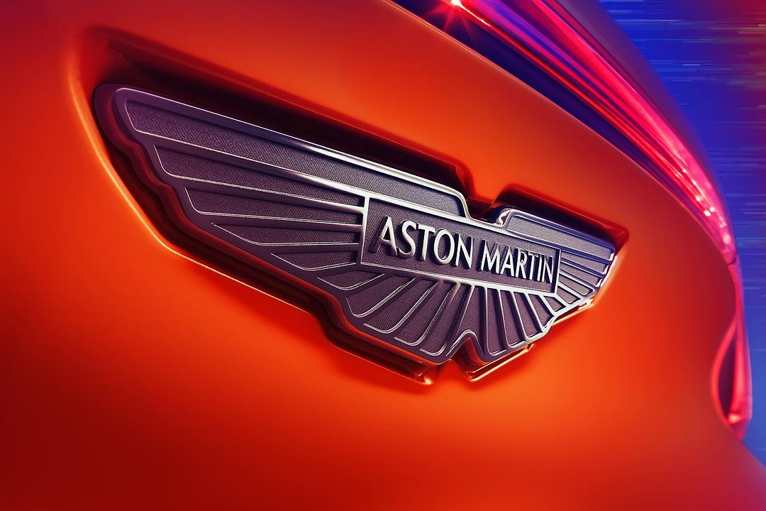 Close-up of the Aston Martin emblem on an orange car body with blurred purple light streaks in the background. Automotive detail shot.