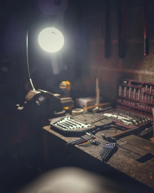 Tools and equipment on a wooden workbench illuminated by a lamp, set in a dimly lit workshop with various instruments.