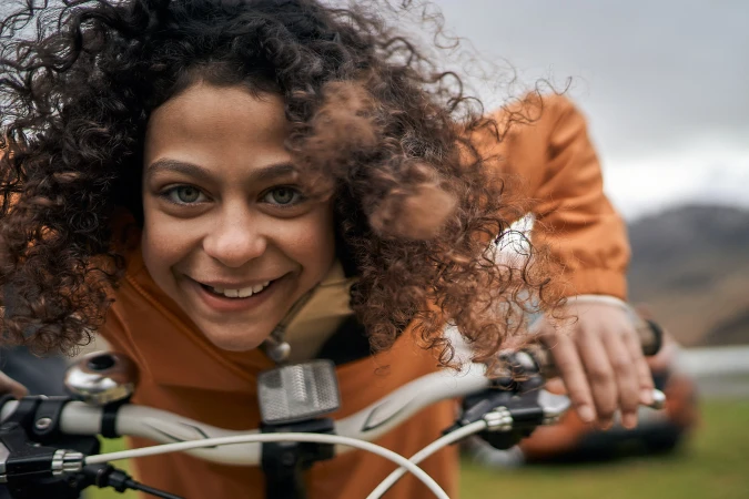 Close-up portrait of a smiling young person with curly brown hair riding a bicycle outdoors in an open landscape