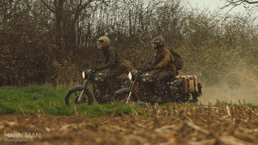 Two motorcycle riders travel on a dirt road through a rural landscape. Both wear helmets and jackets. Trees are visible in the background.