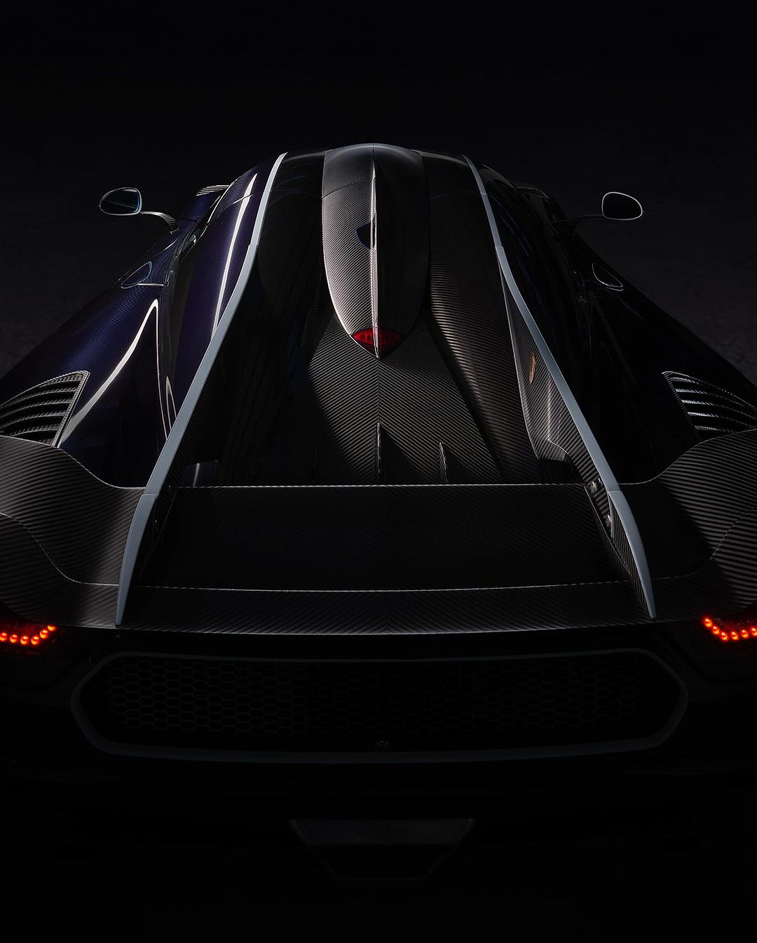 Rear view of a dark blue and black Koenigsegg Jesko sports car with visible carbon fiber detailing in a low-key lighting setup.