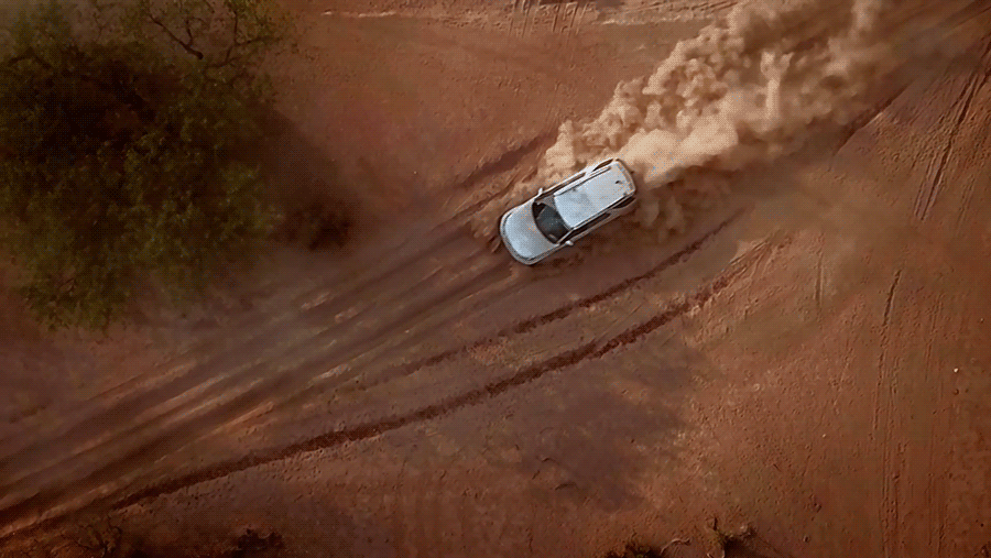 Aerial view of a silver SUV driving on a reddish-brown dirt road, kicking up a cloud of dust, next to green vegetation.