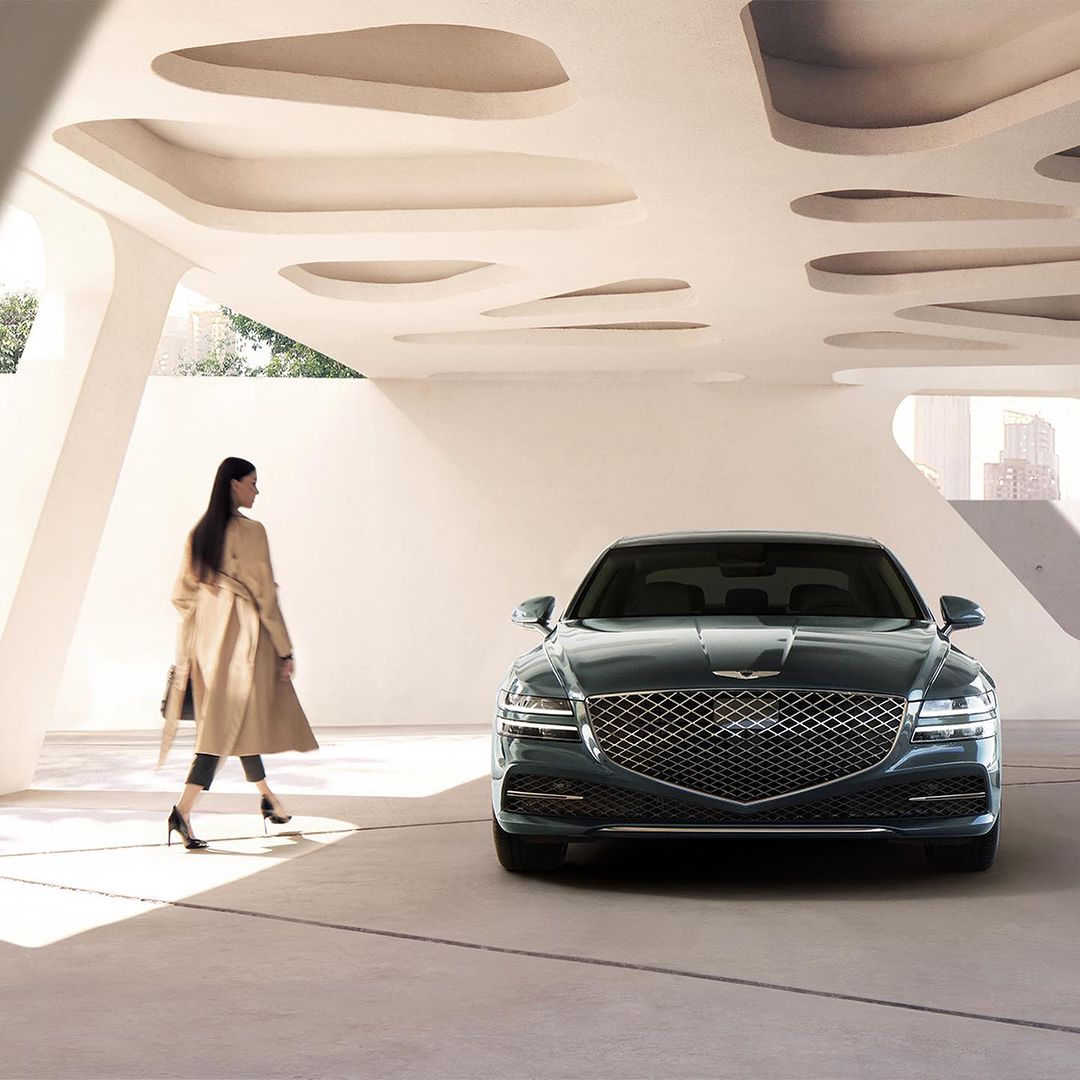 A dark green Genesis sedan parked indoors with an adult woman walking behind it. A modern architectural setting with city skyline.