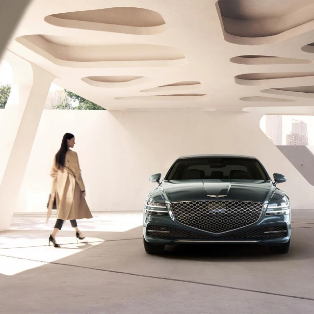 A dark green Genesis sedan parked indoors with an adult woman walking behind it. A modern architectural setting with city skyline.