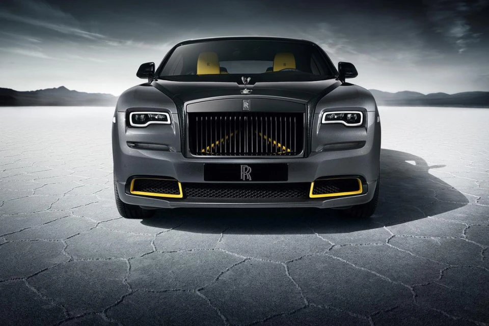 Sleek grey Rolls Royce coupe with yellow trim parked on a cracked salt flat with distant mountains under an overcast sky.