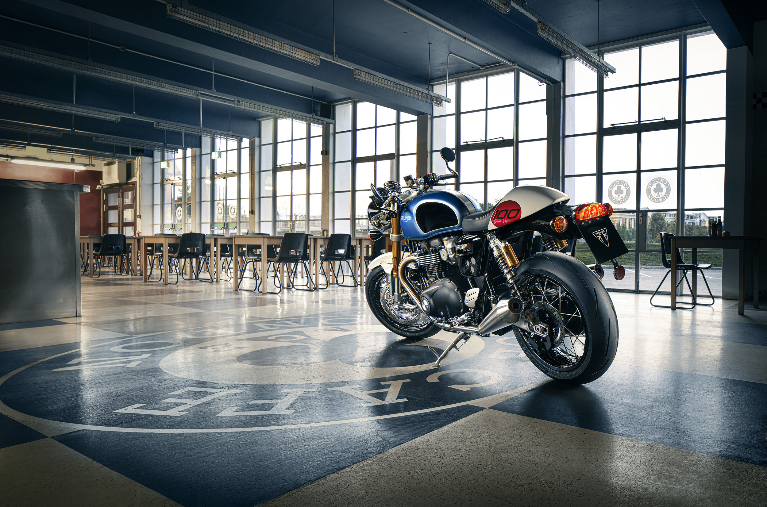 Motorcycle parked inside a high-ceilinged building with large windows, checkered flooring, and tables and chairs.