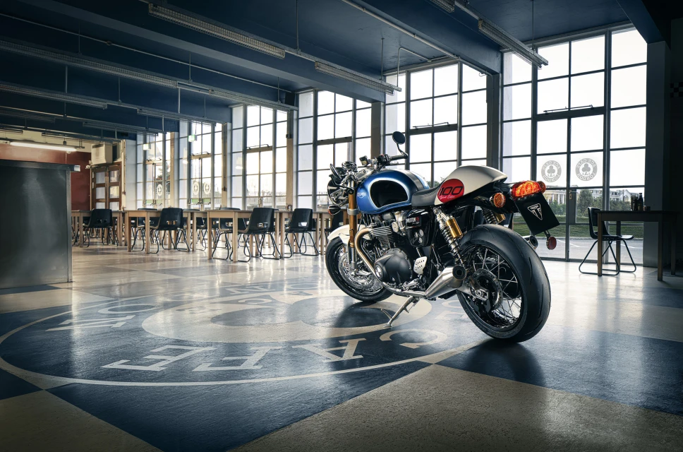 Motorcycle parked inside a high-ceilinged building with large windows, checkered flooring, and tables and chairs.
