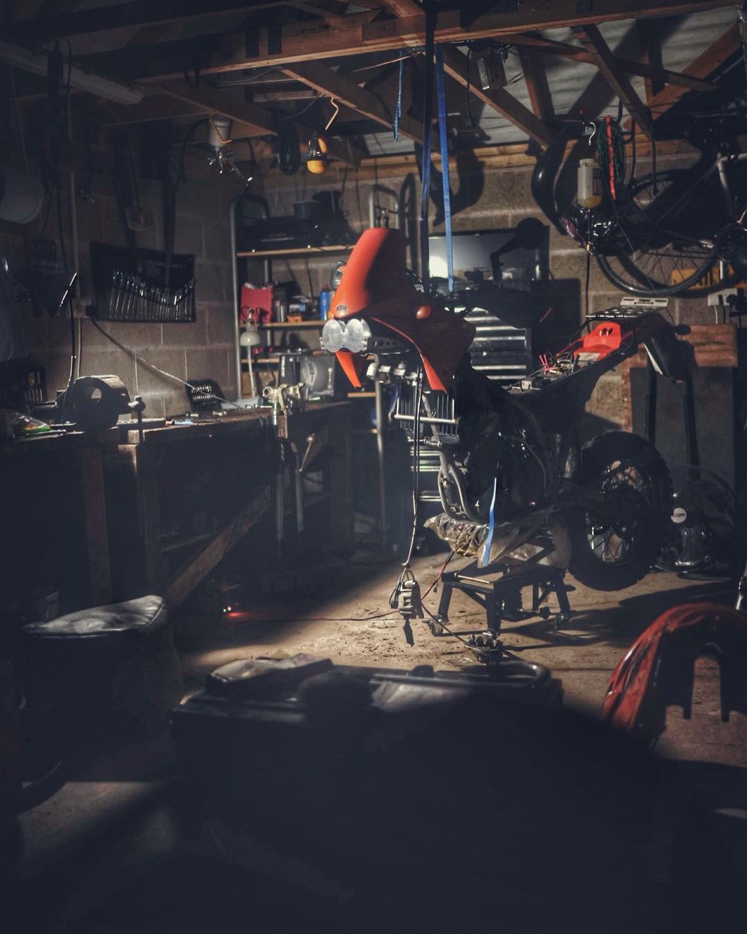 Motorcycle under repair in a cluttered garage. Tools, spare parts, and dim lighting create a workshop atmosphere.