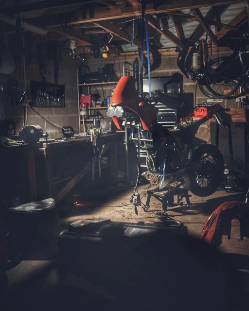 Motorcycle under repair in a cluttered garage. Tools, spare parts, and dim lighting create a workshop atmosphere. 