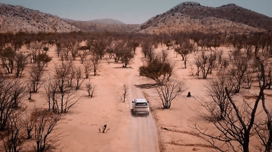 Aerial view of a white SUV driving on a dirt road through a semi-arid landscape with sparse trees and distant mountains under daylight.