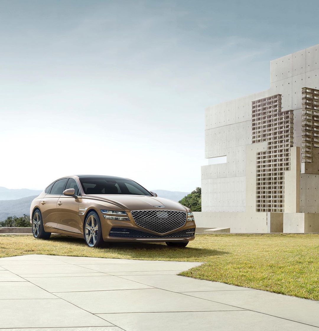 Gold Genesis G80 sedan parked on a paved surface with green lawn and modern architectural building against a blue sky backdrop.