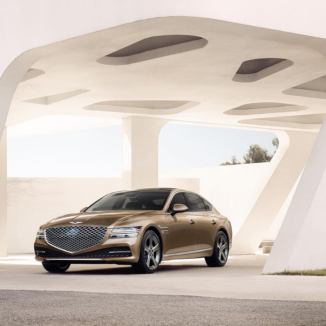 Bronze Genesis G80 luxury sedan parked outdoors, showcasing sleek design, grille, and modern architecture in soft light.