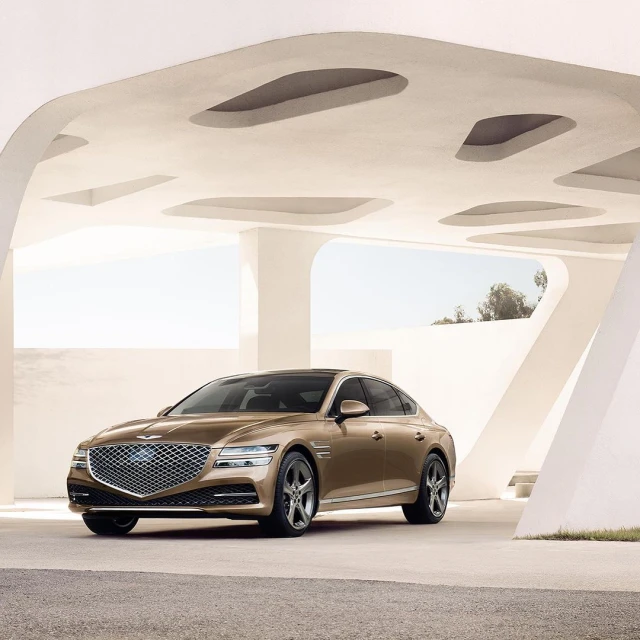 Bronze Genesis G80 luxury sedan parked outdoors, showcasing sleek design, grille, and modern architecture in soft light.
