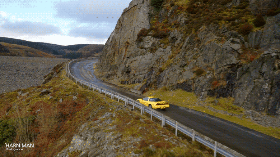Aerial shot of a yellow sports car driving on a wet road next to a rock cliff and mossy hillside, with cloudy sky in the background.