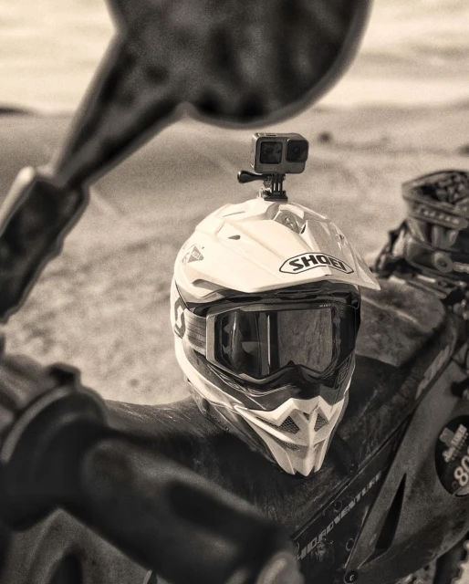 Close-up of a motorcycle helmet with goggles and an action camera attached on top, set against a blurry outdoor backdrop in a sepia tone.