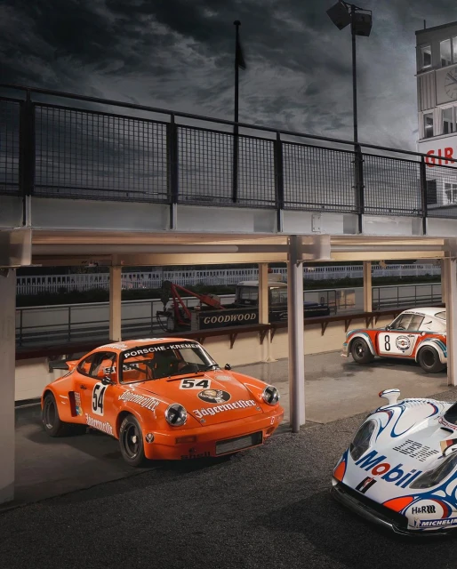 Three Porsche race cars are parked indoors, featuring orange and white exteriors, and a vintage motorsport aesthetic under cloudy skies.