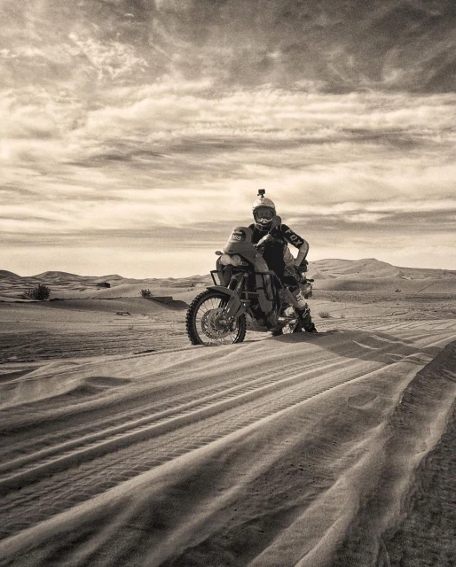 Motorcyclist on an adventure bike in a sandy desert landscape, wearing riding gear and helmet, with a cloudy sky in the background.