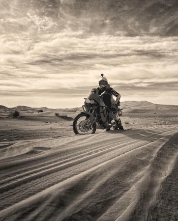 Motorcyclist on an adventure bike in a sandy desert landscape, wearing riding gear and helmet, with a cloudy sky in the background.