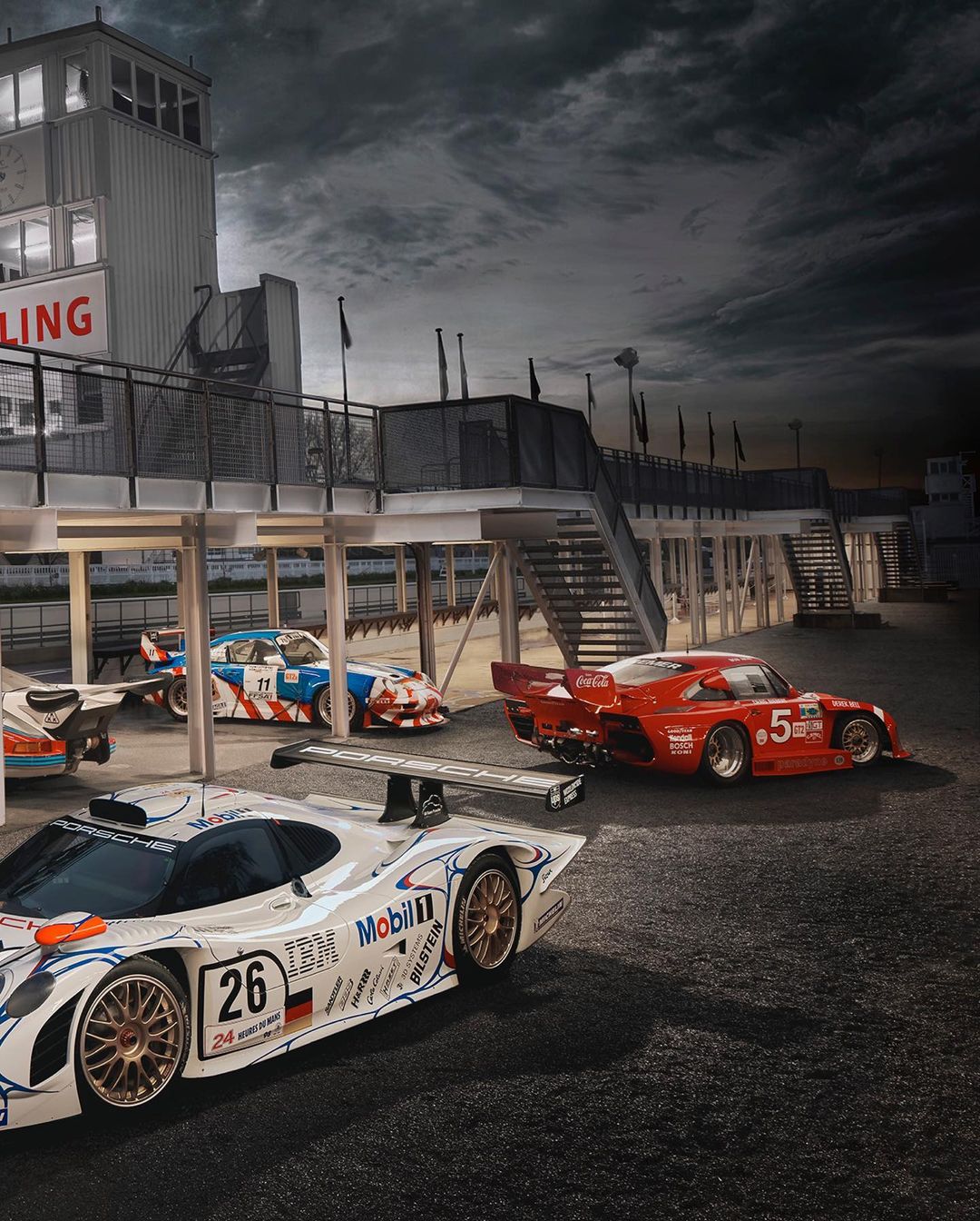 Several racing cars lined up on a track next to a pit lane, shot on a cloudy day, showcasing speed and automotive design.