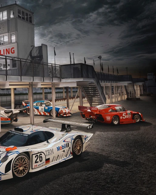 Several racing cars lined up on a track next to a pit lane, shot on a cloudy day, showcasing speed and automotive design.