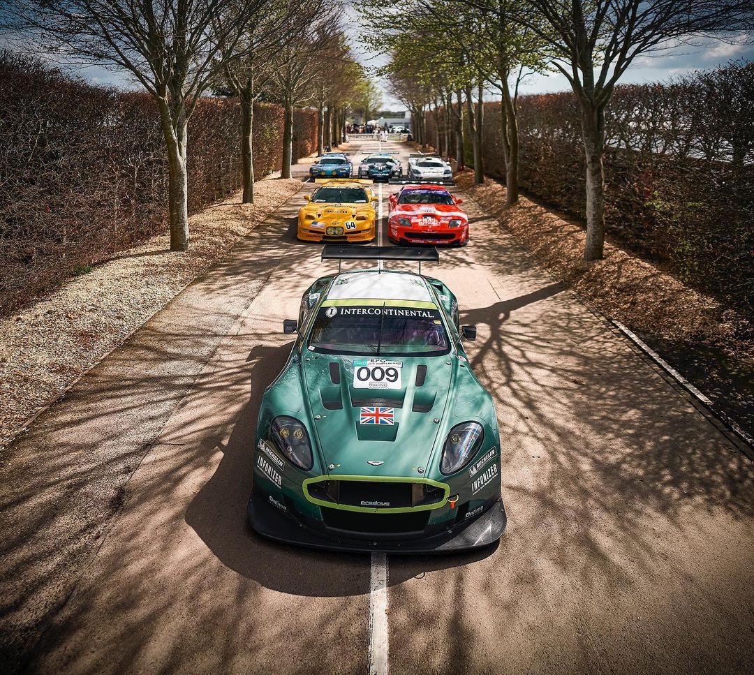A line of classic race cars in green, yellow, and red leading the pack on a tree-lined track, captured in bright outdoor lighting.