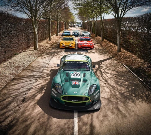 A line of classic race cars in green, yellow, and red leading the pack on a tree-lined track, captured in bright outdoor lighting.