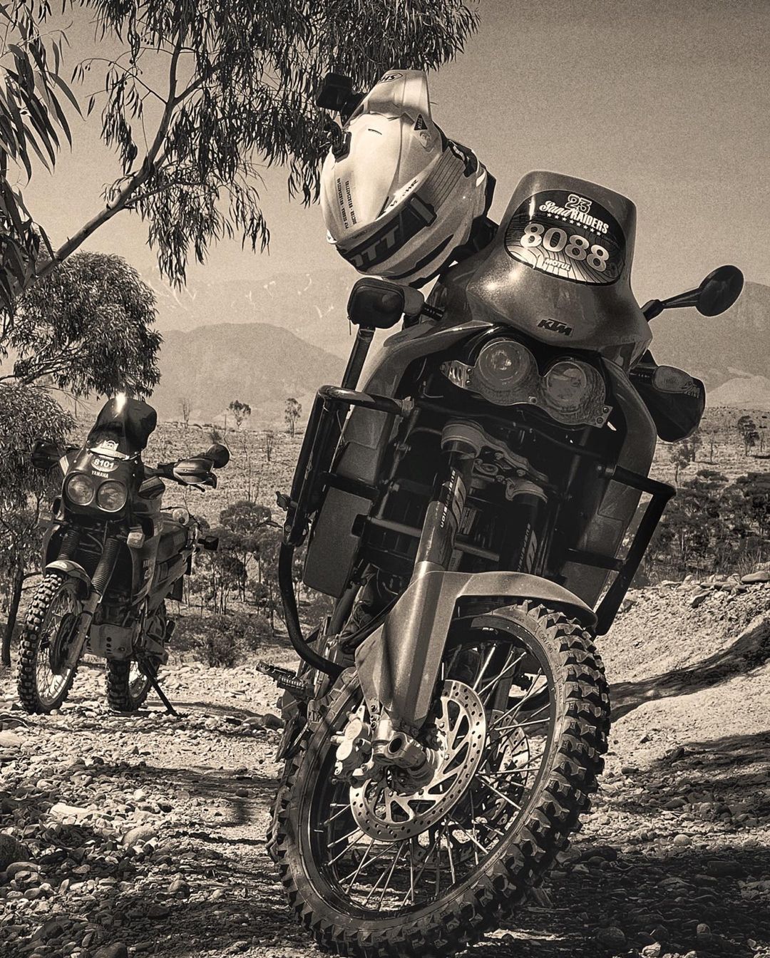 Two enduro motorcycles parked on a rocky, barren landscape under a clear sky, one with a helmet resting on the front, black and white.