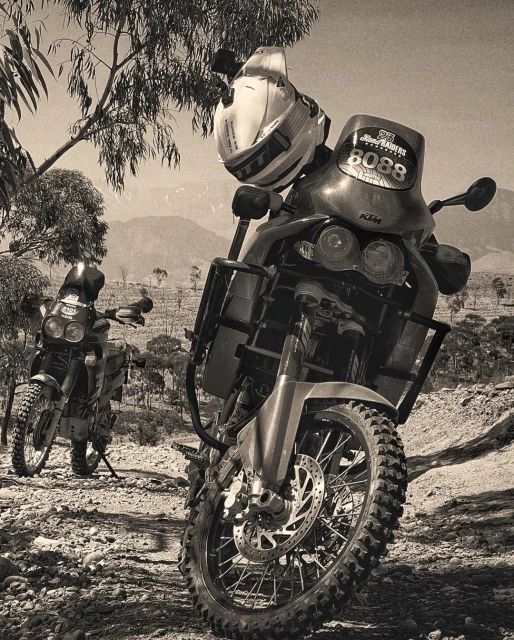 Two enduro motorcycles parked on a rocky, barren landscape under a clear sky, one with a helmet resting on the front, black and white.