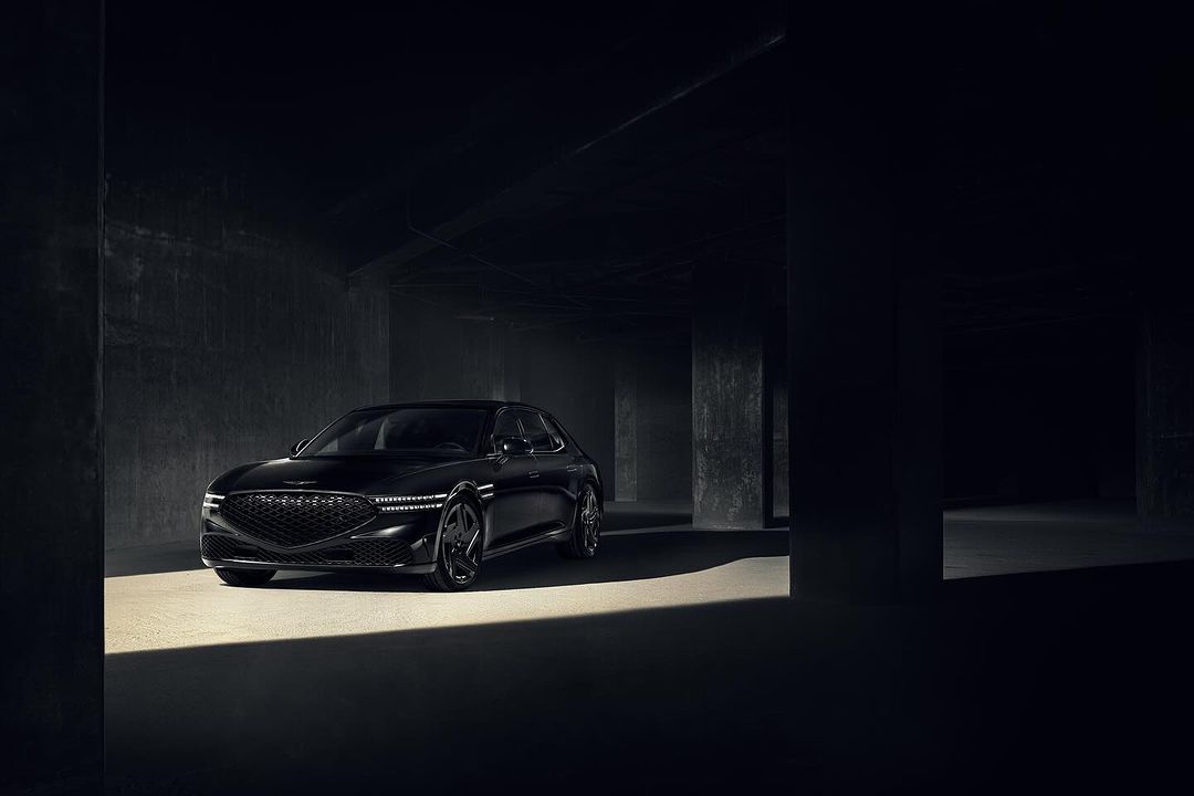 Black Genesis G80 sedan parked in a dimly lit concrete parking garage with light reflecting off the front hood, showcasing automotive design.