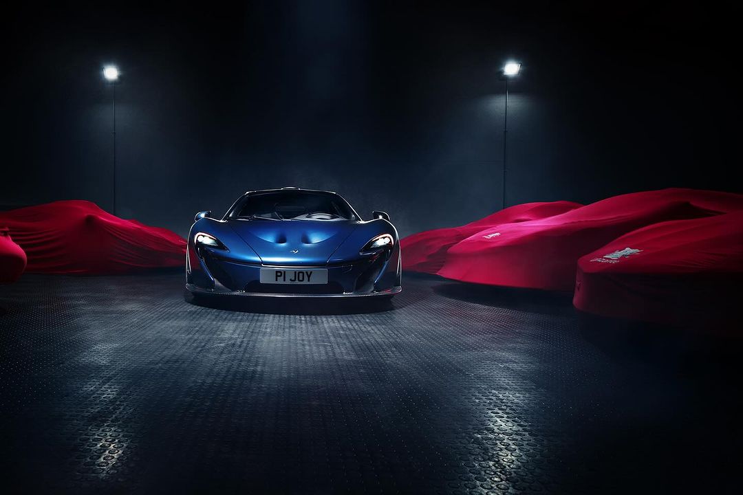 Bright blue McLaren P1 JOY car on a dark, shiny platform in a showroom with other covered sports cars lit by overhead lighting.