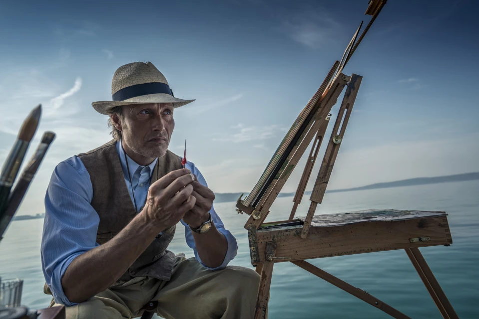 Man in straw hat sits outdoors by water painting on easel, holding a small brush, dressed in light blue shirt and brown vest.