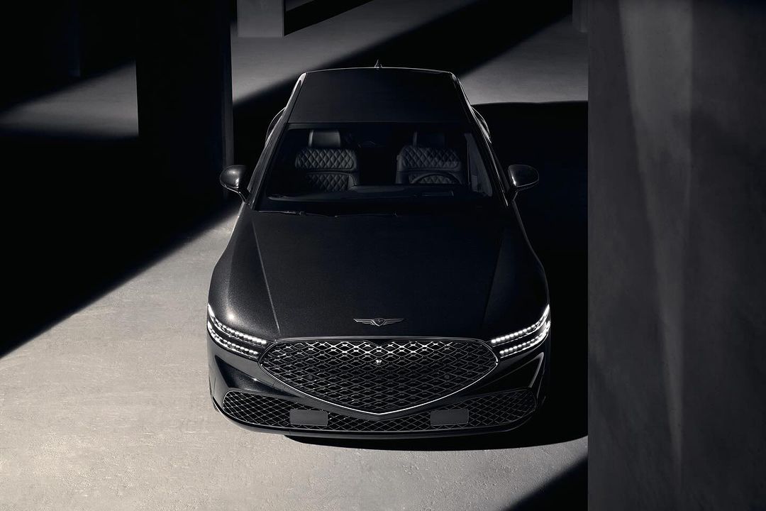 Front view of a dark gray Genesis sedan parked indoors, showcasing its grill, headlights, hood, and sleek design in a low-key lighting environment.