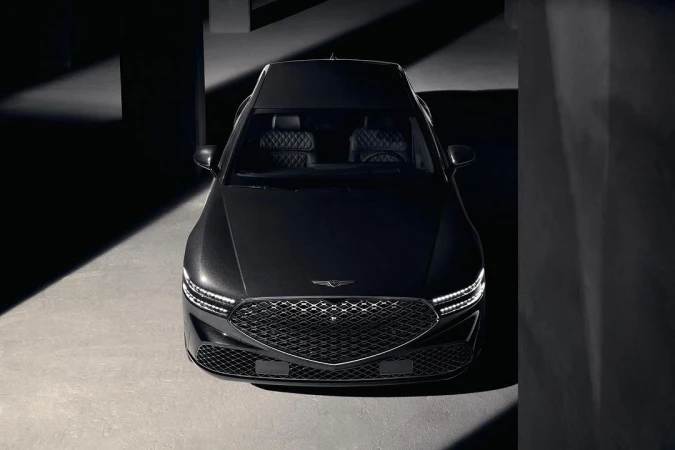 Front view of a dark gray Genesis sedan parked indoors, showcasing its grill, headlights, hood, and sleek design in a low-key lighting environment.