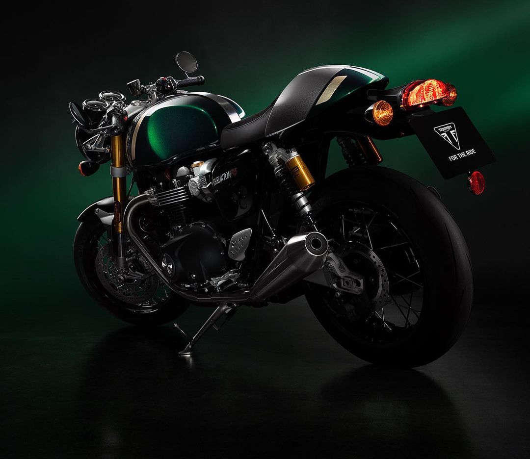 Rear view of a Triumph Thruxton RS motorcycle featuring a dark green fuel tank, black seat, and visible taillight on a dark background.